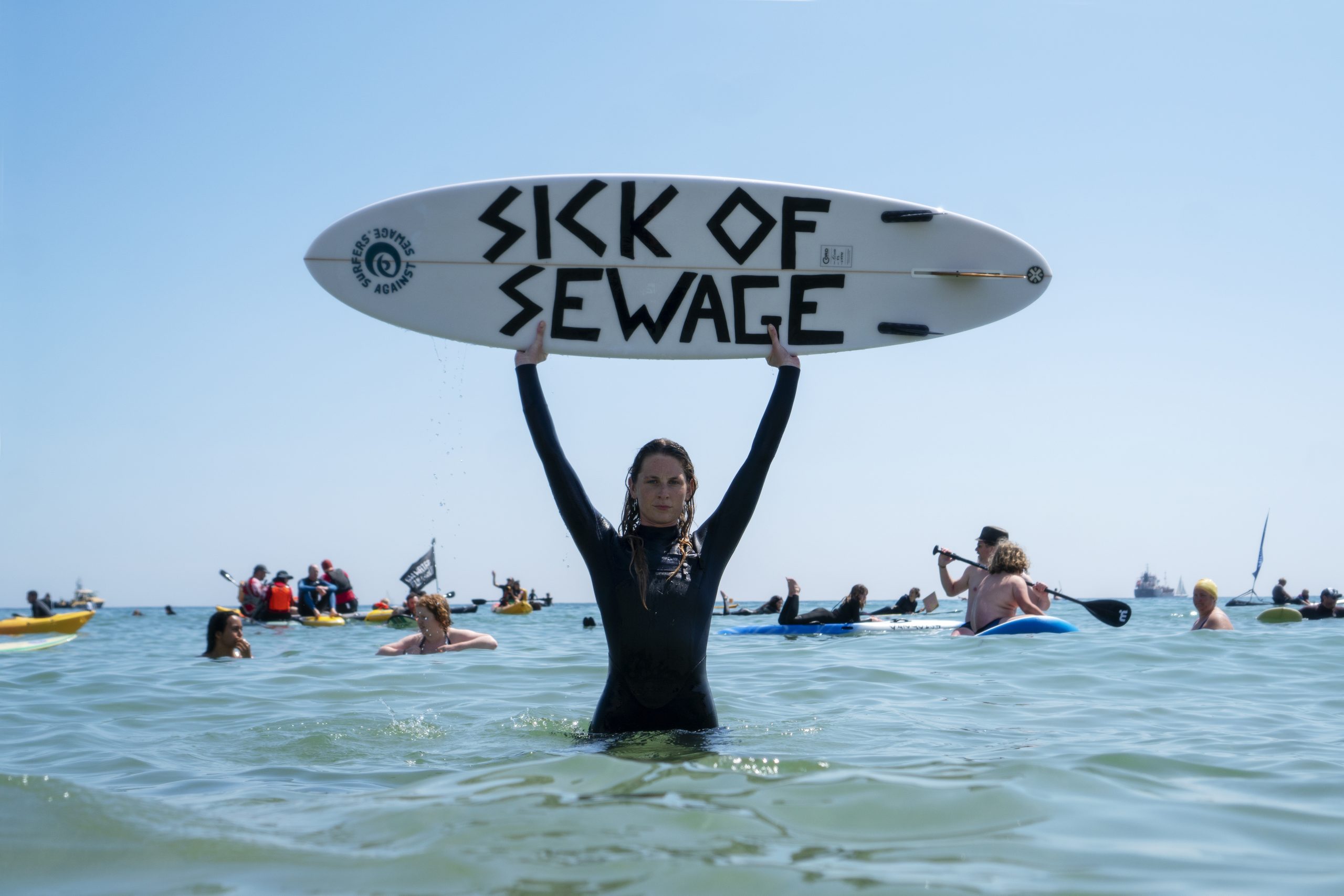 A Surfer in the sea holding a surfboard with the words "Sick of Sewage" written on it